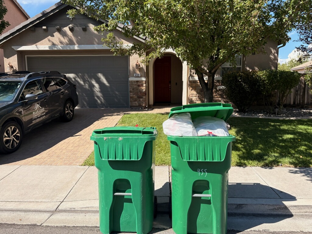 Two green trash bins at a residential curb, one with bags extending above its open lid